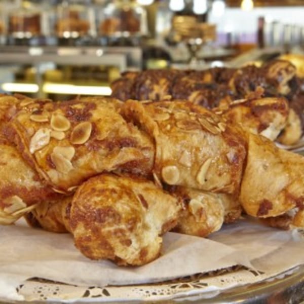 Golden brown almond croissants on a white paper doily, with other pastries blurred in the background.