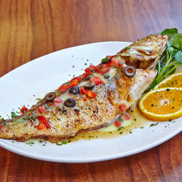 Whole grilled fish topped with diced tomatoes, olives, capers, and a lemon sauce, garnished with orange slices.